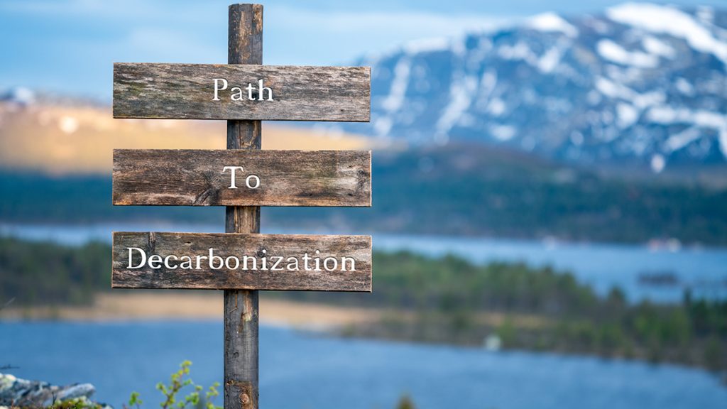 Wooden sign that reads "Path to Decarbonization." In the background and out of focus is a lake and a snowy mountain. 