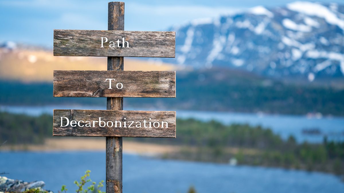 Wooden sign that reads "Path to Decarbonization." In the background and out of focus is a lake and a snowy mountain.
