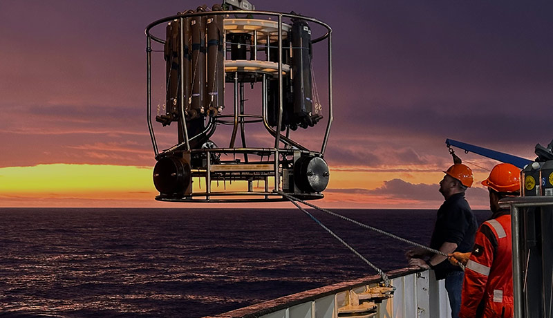 Researchers dipping sensing instrument into water during sunrise.