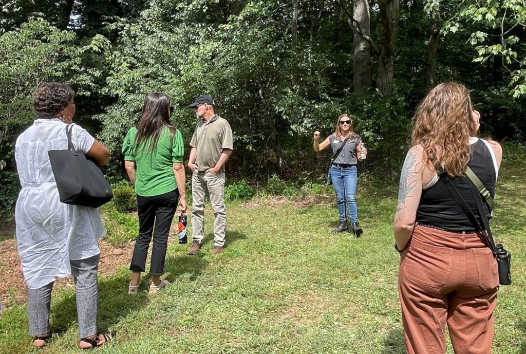 In Rutgers Gardens, the attendees stand on the grass in front of a forest listening to a tour guide talk