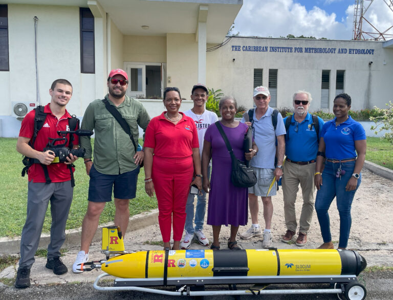 Ben Lerner holding a camera and posing with others in front of the The Caribbean Institute for Meteorology and Hydrology