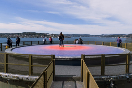 Flat, colorful, circular art installation surrounded by a fence overlooking water