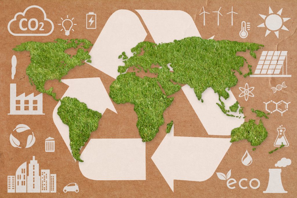 A flat, green, grass textured world map against a cardboard background with various symbols relating to sustainability behind and surrounding it such as recycling arrows and solar panels