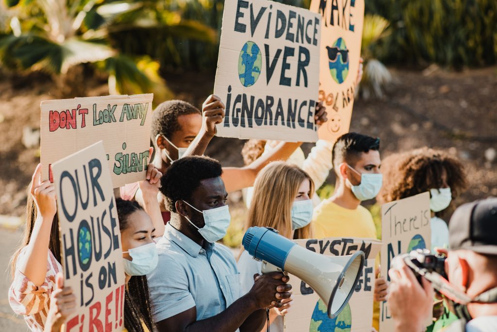Young multiracial demonstrators protesting for climate change while wearing safety masks outdoor in the city, holding signs like "Evidence Over Ignorance"