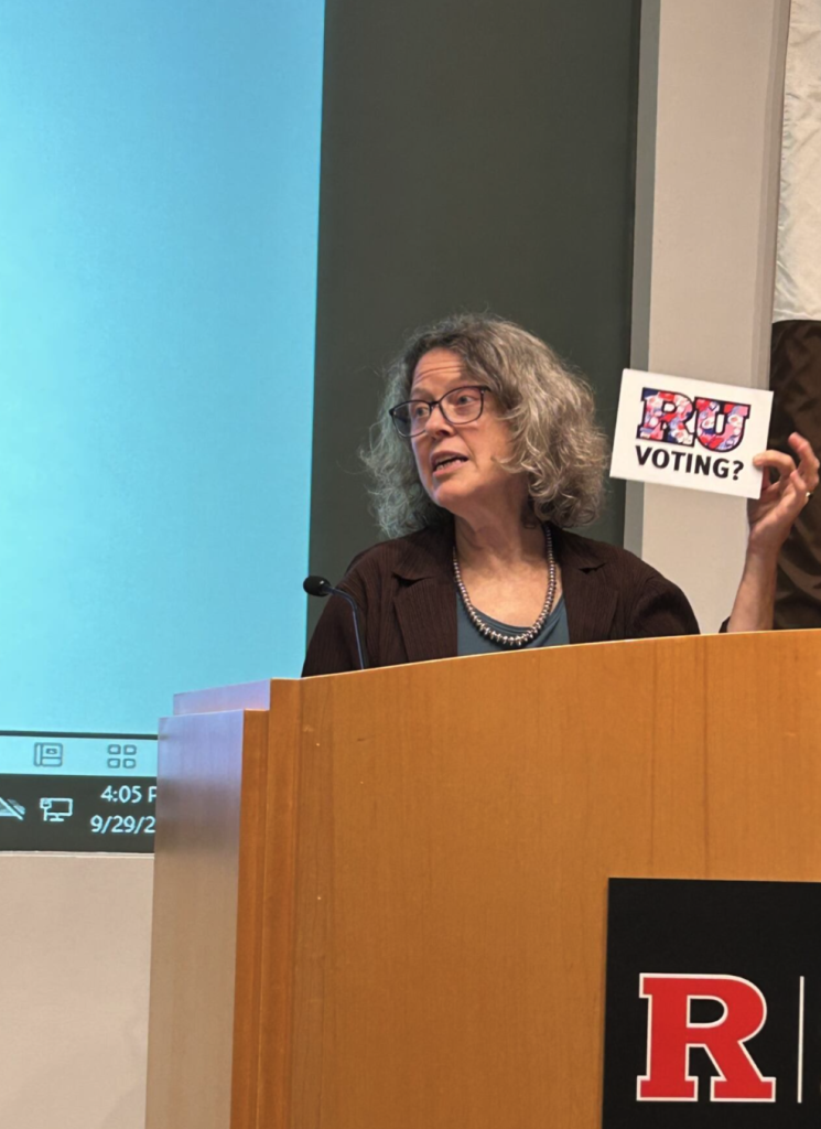 Professor Cymie Payne speaks at a podium, holding up an "RU Voting?" card