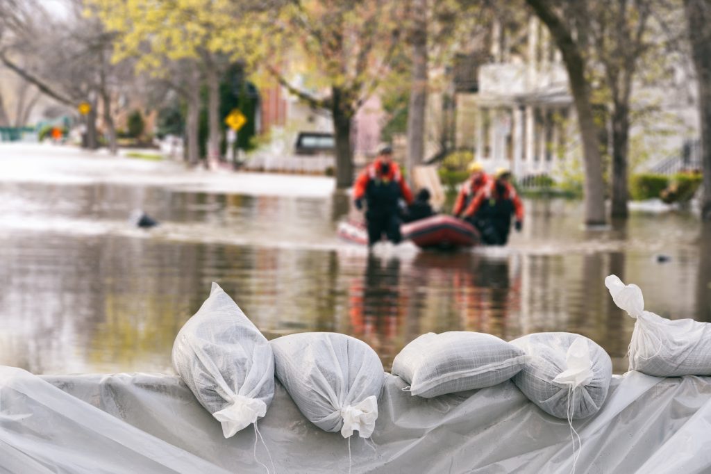 Flood Protection Sandbags in the front with flooded homes in the background on a tree lined street. A lifeboat pushed by safety workers moves through the water.