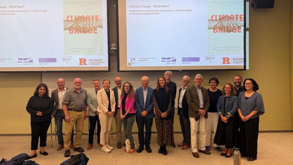 Panel speakers standing together on stage in front of a big screen with the Climate Bridge poster on it and the words "Climate Change – What Now?"