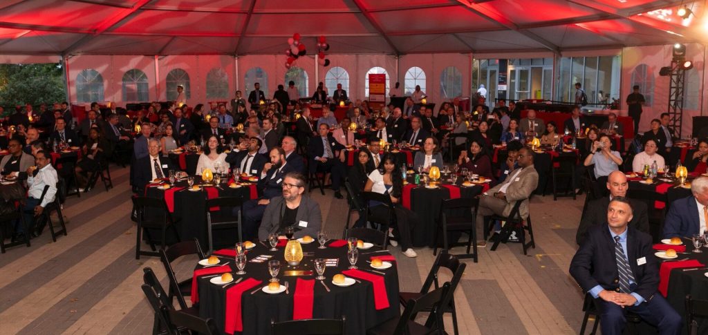 Underneath a very large white tent, circular tables with formally dressed attendees sitting around them, plates and silverware on top of them 