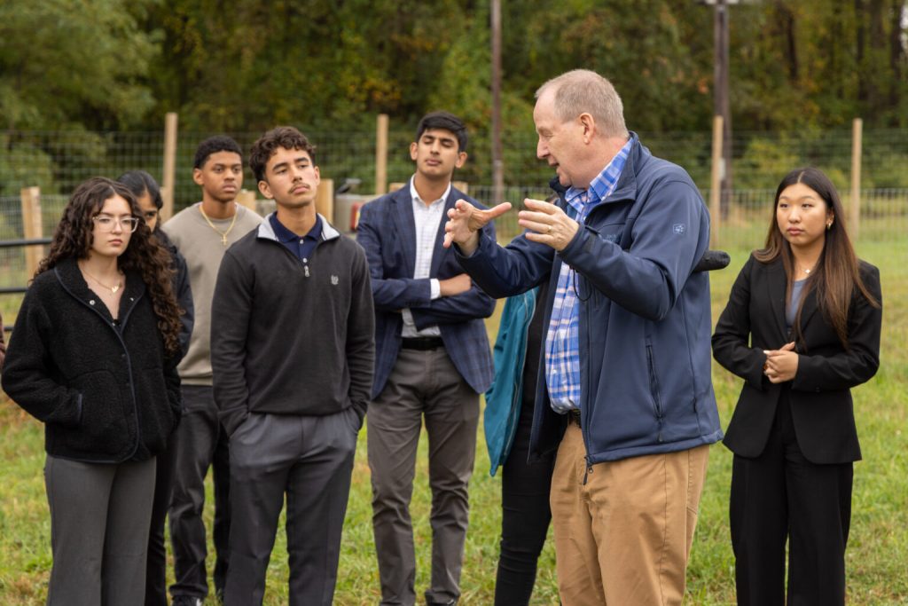 AJ Both stands in front of a small group of attentive students, outside on the grass with a fence and trees in the background
