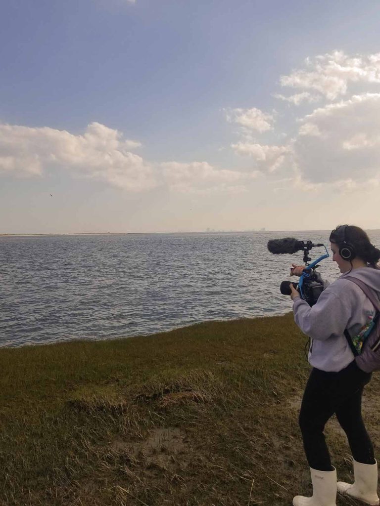 Kimberly Hansen holding a video camera with an attached microphone, standing on the marshland in front of the waterline, videoing the water