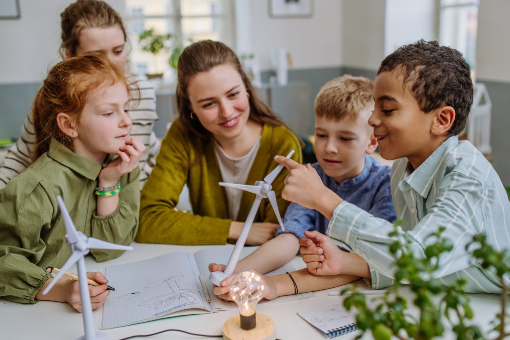 Young teacher with model of wind turbine sitting at a desk with three curious pupils, teaching them about about wind energy.