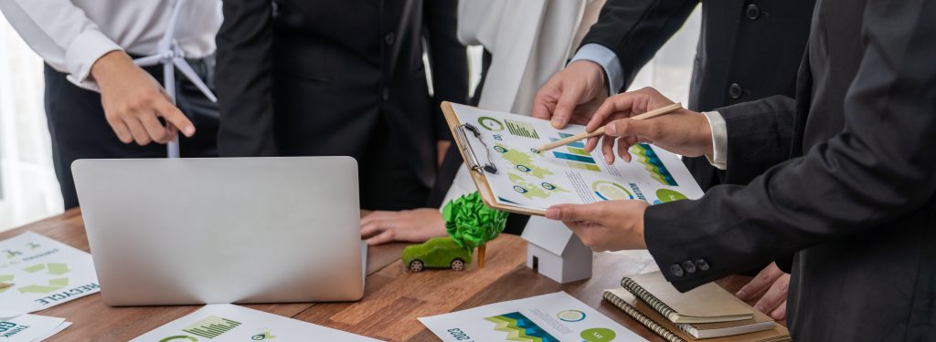 Professionally dressed group of business men/women meeting using laptop, charts on a clipboard, and physical models to plan strategy and discuss marketing of eco-friendly and clean energy products. 