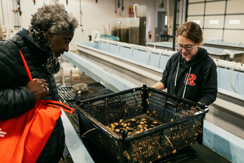 Sophia Piper showing a curious participant a black crate full of clams in a long chute of water 