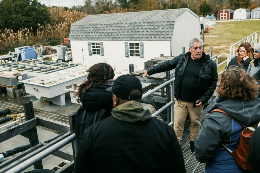 Michael De Luca talking and gesturing to a group of people as they stand looking down at a white building, the oyster nursery