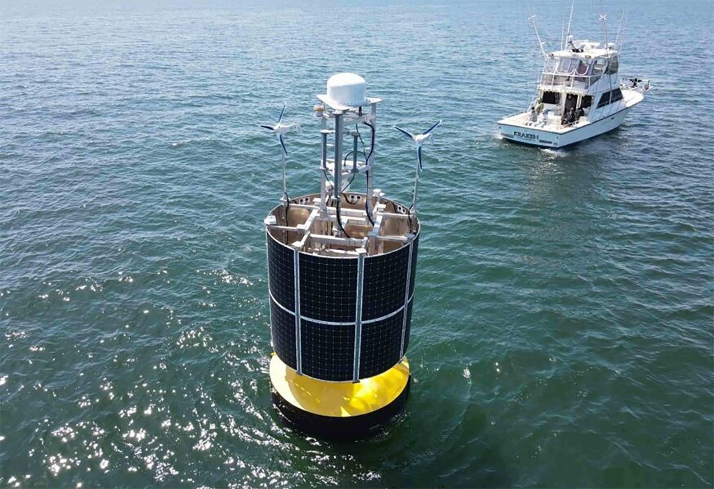 View from above of a large buoy wrapped in solar panels floating in the middle of the ocean, a small boat off to the side with scientists watching.
