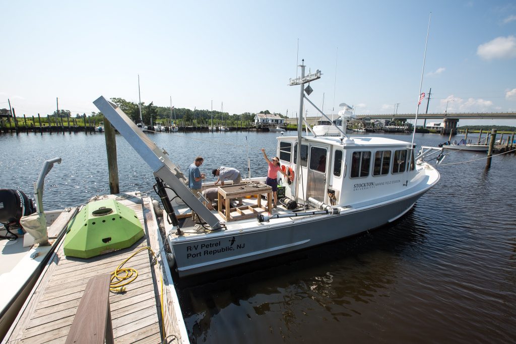 A dock with a small boat, labelled "Stockton", just off the side. Scientists conduct experiments on the boat's deck.