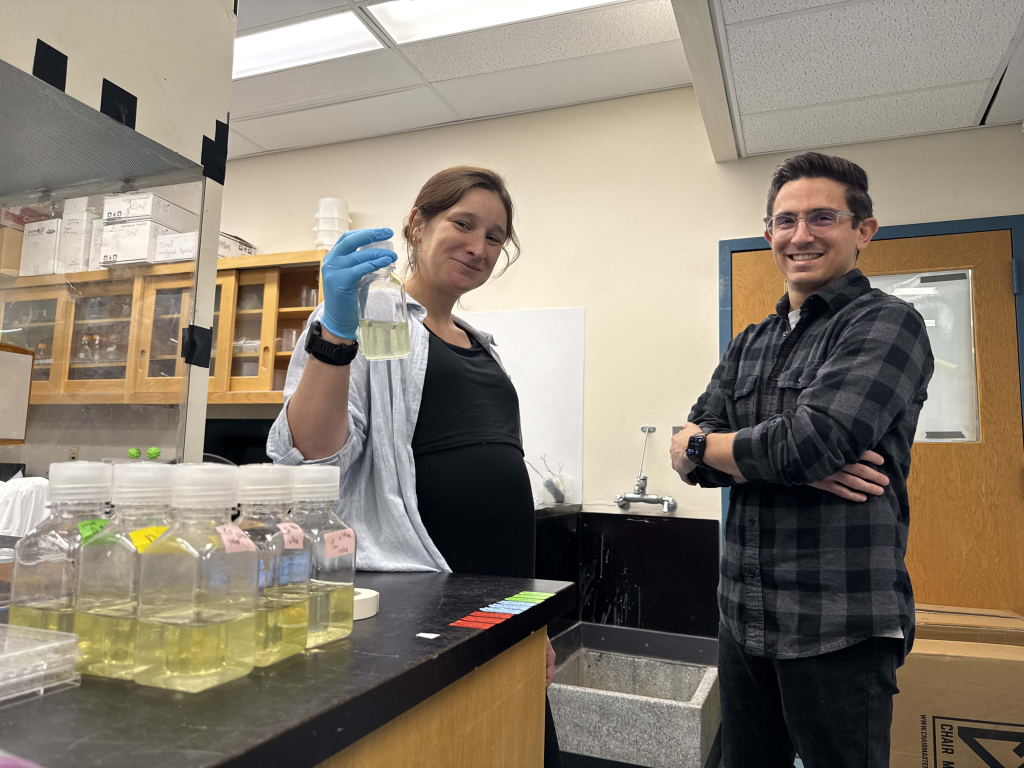 Corday Seldon and Joe Gradone stand side by side in a wet lab, Seldon holding up a bottle filled with pale yellow liquid, clearly part of some experiment, while Gradone crosses his arms with a smile.
