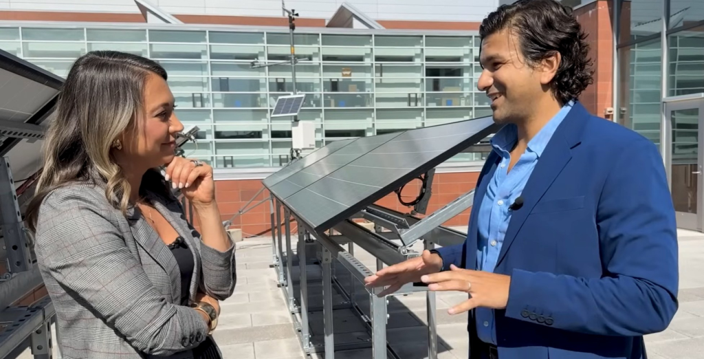Aziz Ezzat, standing on the right, gestures with his arms as he talks animatedly to Ginger Zee, standing on the left. They are outside, talking in front of a row of solar panels.