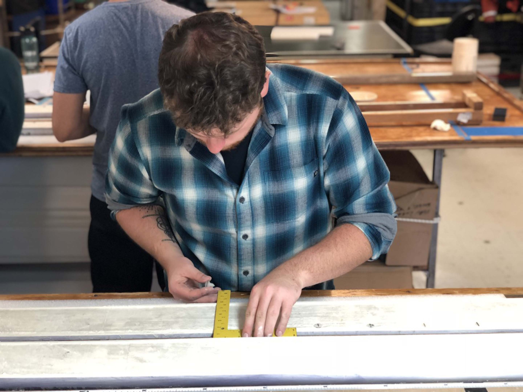 Ryan Glaubke leans his head down to concentrate on using a measuring tool against a metal item in a lab room, where other students are working behind him.