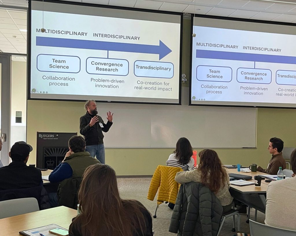 Professor Kay Bidle stands at the front of the room giving a speech with slides projected in the background. Other people sit at tables watching him.