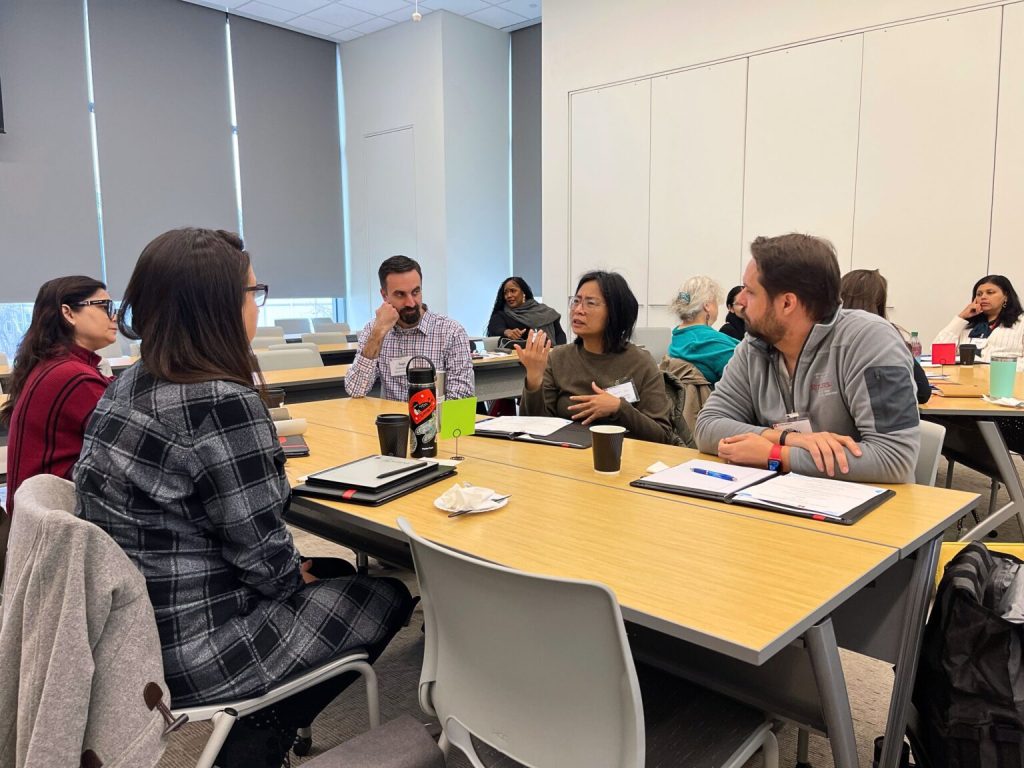 5 attendees sit at a table talking to each other. In the background, others are doing the same at other tables.