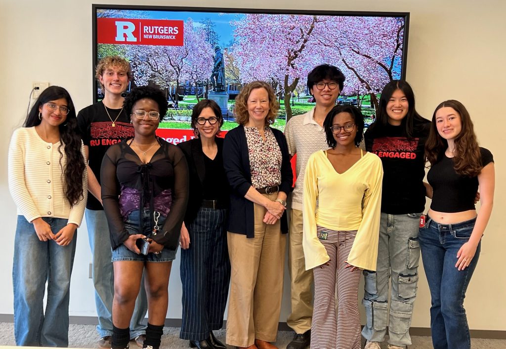 The mentioned individuals standing together and smiling for the photo in front of a projector with an image of Rutgers campus on it