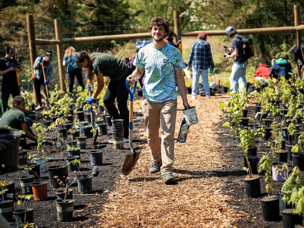 Josh Kover, holding a shovel and small bucket, walks down a path. On either side are many small saplings in pots on fertile soil and community participants planting them into the ground.