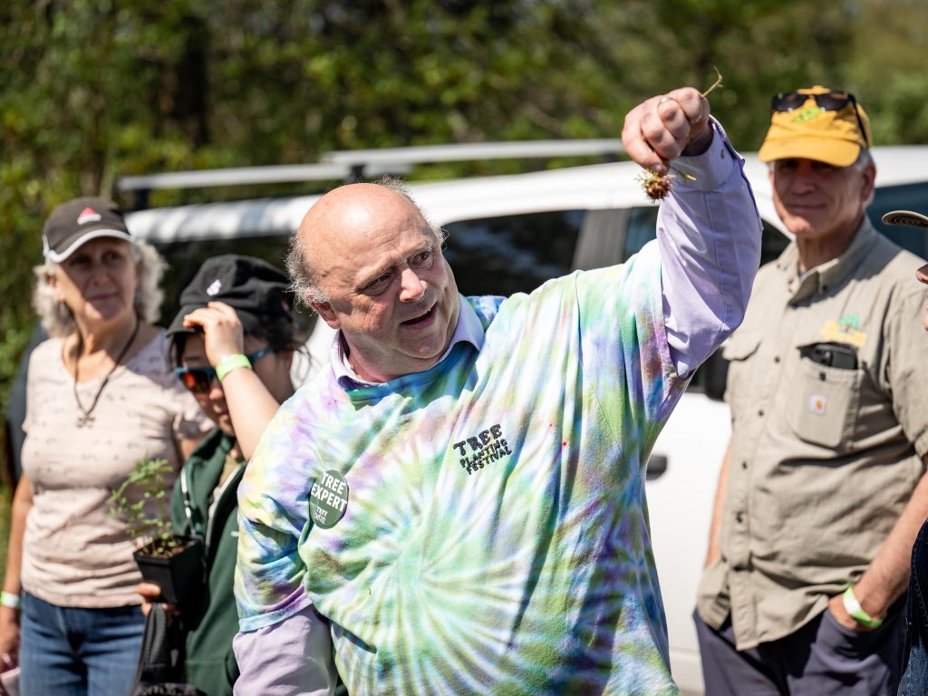 Dr. Jason Grabosky holds up a pulled-up plant high in demonstration for the onlookers to see as he gives his speech. Behind him a couple participants can be seen, as well as a white van.