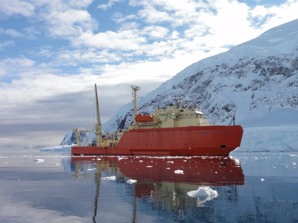 A large boat, a research vessel, floats between bits of ice on crystal clear water, a snow covered mound of land in the background