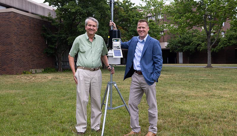 David Robinson and John Krasting stand on either side of the weather station they set up, sensing equipment attached to the middle of a pole set to the ground with a tripod