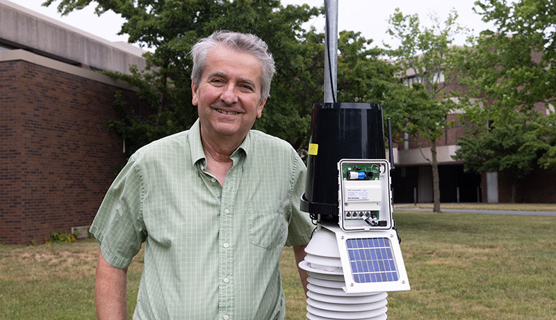 David Robinson stands smiling next to the technological headpiece of the weather station, holding the pole
