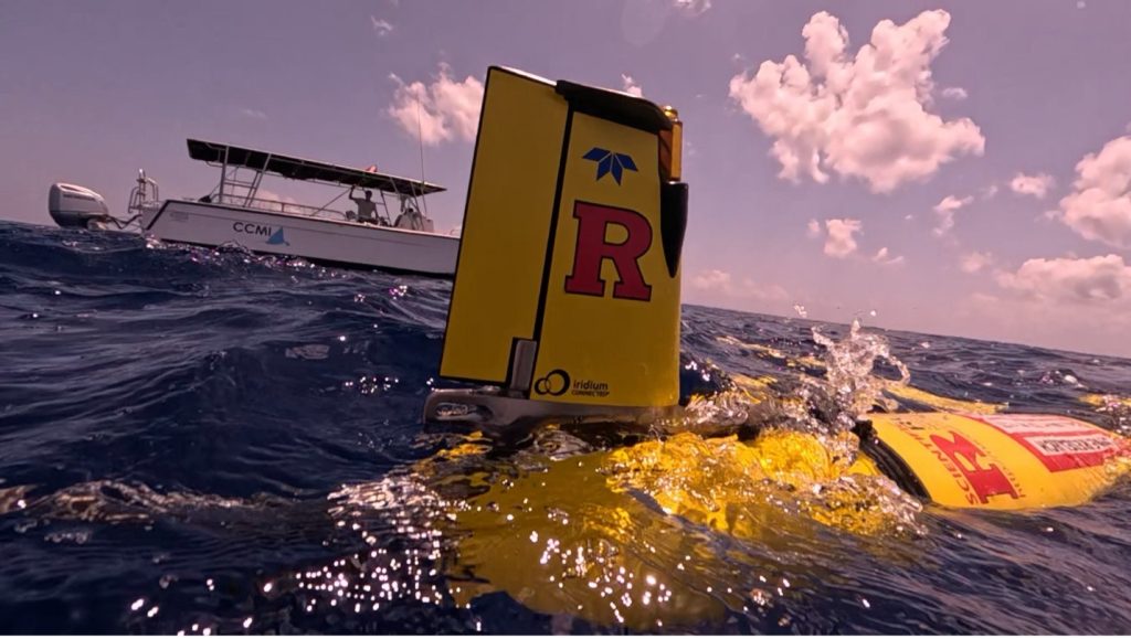 RU37 glider half submerged in the waves, its hull peaking up from the ocean, with researchers watching from a small boat in the background
