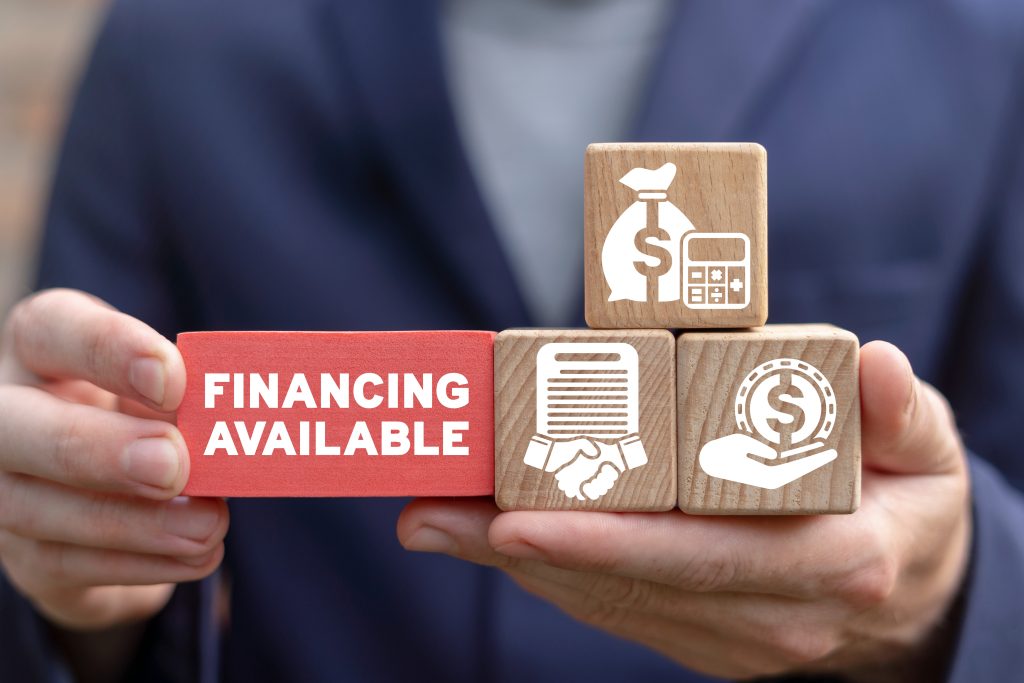 Hands holding wooden blocks with icons representing funding, partnerships, and financial support next to a red block labeled “Financing Available.”