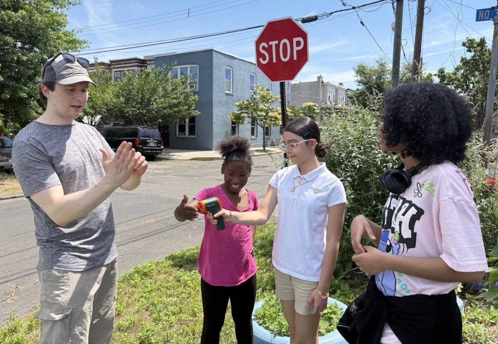Standing outside in an area of grass and bushes on a sunny day, James Shope gestures with his hands as he explains surface temperature measurement to three young students standing on the right, one of whom is holding a measurement tool. 