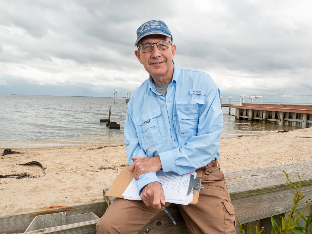 Kenneth Miller sits with a clipboard, papers, and pen on a wooden fence in front of the shore. The waves, sand, and a dock are behind him.