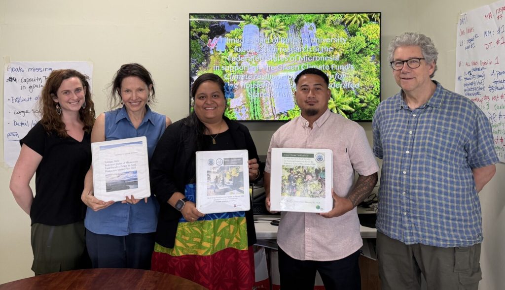 Five individuals, including Dena Seidel and Jim Simon, stand in a meeting room in front of a small presentation screen that projects a colorful slideshow. Three individuals hold up binders with cover graphics representing policies and plans.