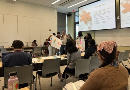 A group of participants stand in the front of a classroom presenting a slideshow on the big screen and holding up posters. Onlookers sit at long tables and listen.