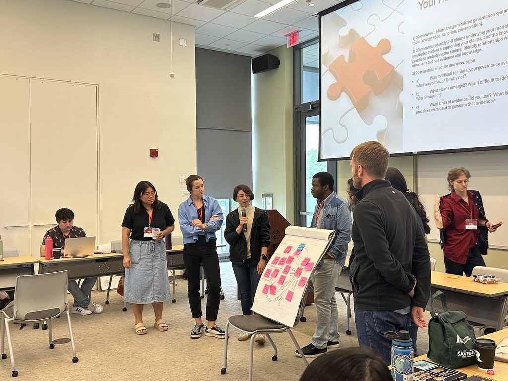 Closer shot of a group of presenting individuals with a slideshow in the background and a propped-up poster with a handmade concept map on display. One of the participants is speaking into a microphone.