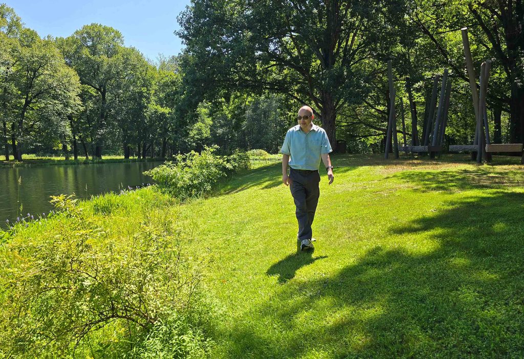 Qizhong "George" Guo walks on the grass next to a large body of water, trees in the background, sunny day.