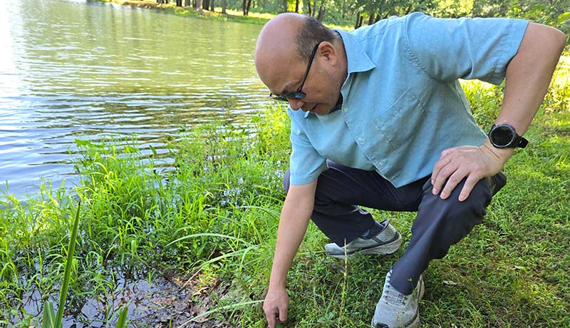 Qizhong "George" Guo crouches down on the grassy bank surrounding a body of water. He extends one hand to the edge where the water and ground meet.