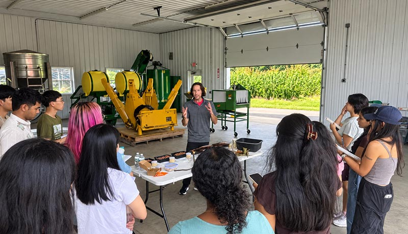Thomas Molnar lectures to a group of students who stand around him and his demonstration table. Upon the table are plastic boxes of hazelnuts. They all stand in some sort of warehouse/garage with machinery in the background.